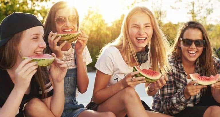 A group of smiling young people sit together outside, enjoying slices of watermelon. The scene is lit by warm sunlight, suggesting a summer afternoon gathering. Casual clothing, sunglasses, and headphones add to the relaxed, youthful vibe. The overall mood is cheerful and carefree, with friends sharing a fun moment.