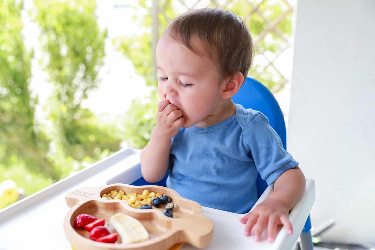 A young toddler sits in a high chair, eating snacks with one hand. The child wears a light blue shirt and is focused on the food. A wooden divided plate holds sliced strawberries, banana pieces, blueberries, and small cereal or corn puffs. The scene is brightly lit with a soft, green outdoor background visible behind the child.